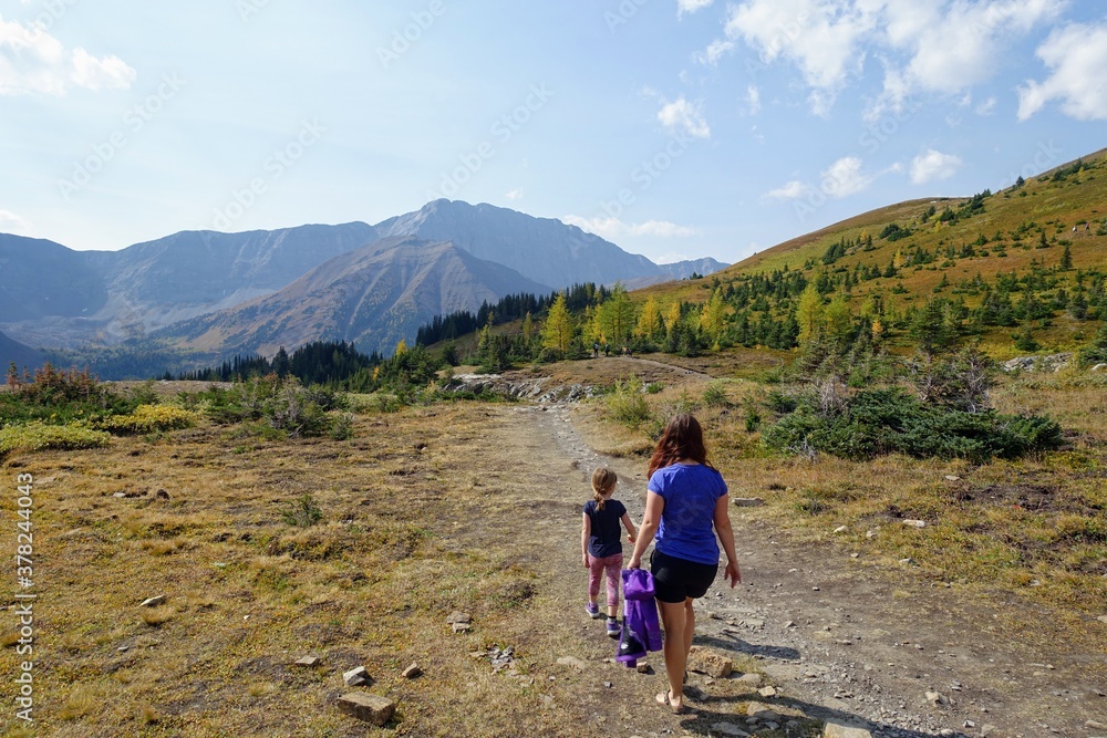 A mother and daugher hiking a beautiful trail above the treeline with a huge mountain in the background during a sunny day in autumn, along the Ptarmigan Cirque trail in Kananaskis, Alberta, Canada