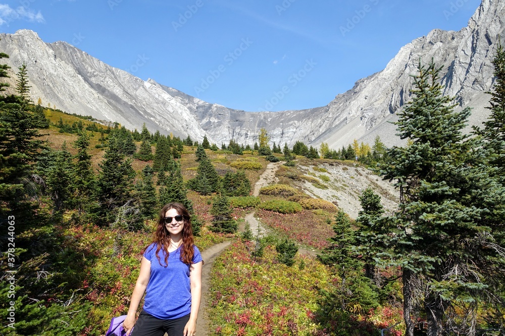 A young pretty woman smiling along a hiking trail with a huge mountain in the background during a sunny day in autumn, along the Ptarmigan Cirque trail in Kananaskis, Alberta, Canada