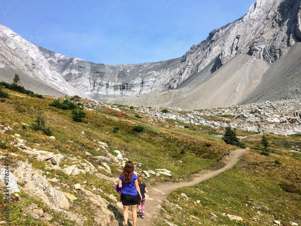 A mother and daugher hiking a beautiful trail above the treeline with a huge mountain in the background during a sunny day in autumn, along the Ptarmigan Cirque trail in Kananaskis, Alberta, Canada