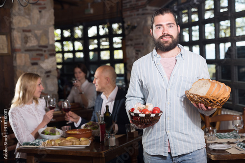 Hospitable young owner of cosy country restaurant meeting guests with traditional meals in hands