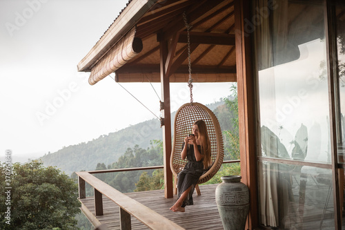 Tourist woman swing on wicker rattan hang chair in the jungle, nature mountains view, hold in hands cup of tea/coffee © Yevhenii