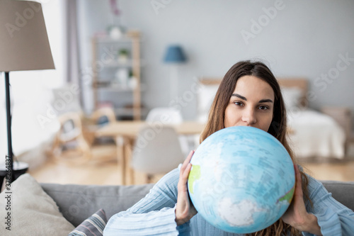 Smiling young woman holding earth globe sitting on the couch at home. Planning vacation.