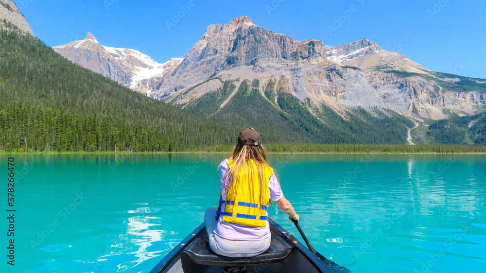 Emerald Lake Canoeing. The view on the back of woman in yellow life jacket paddling from canoe on the beautiful turquoise lake. The mountain and forest in the background. Emerald Lake Canoeing. The view on the back of woman in yellow life jacket paddling from canoe on the beautiful turquoise lake. The mountain and forest in the background.