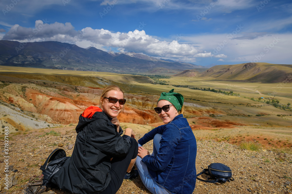 Two young girls in sunglasses sitting on hill look at camera and smile. Women travelers are photographed against the beautiful mountains on sunny day. Active recreation and adventure. Two young girls in sunglasses sitting on hill look at camera and smile. Women travelers are photographed against the beautiful mountains on sunny day. Active recreation and adventure.