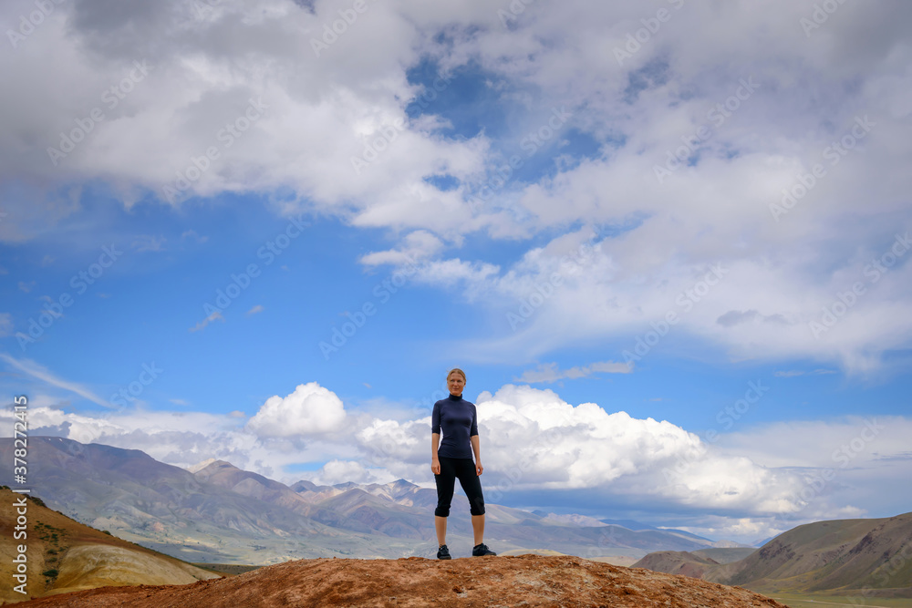 Young happy woman of athletic build in tight clothing stands on the top of mountain against the blue sky on sunny day. Concept of a healthy lifestyle, active recreation, success, travel.