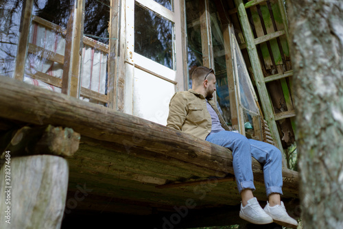 Stylish man sitting on terrace of wooden country retro house surrounded by trees. Enjoy isolation.