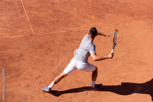 Professional tennis player performs forehand hit on clay tennis court. Young male athlete with tennis racket in action. Junior tennis sport. Back view, shadow, copy space © Elena