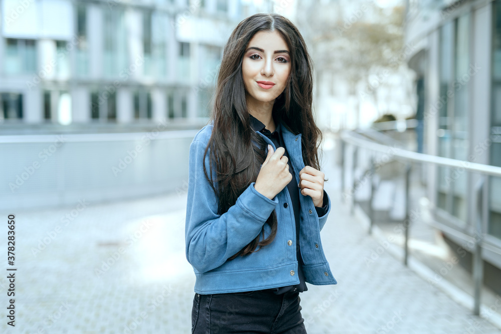 teenage girl posing in city while look neutral into camera with make up and hair done and hands on collar of blue jacket