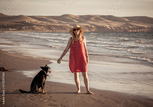Happy attractive mature woman with her pet walking on friendly dog beach at sunset