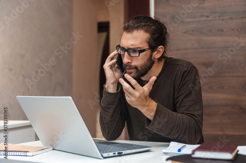 Portrait of serious man with beard talking on mobile phone and working at laptop at home