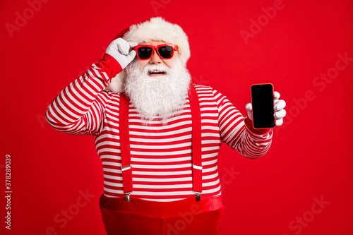 Portrait of his he nice handsome cheerful glad bearded fat Santa holding in hand gadget 5g app roaming service sale order delivery shopping isolated on bright vivid shine vibrant red color background © deagreez
