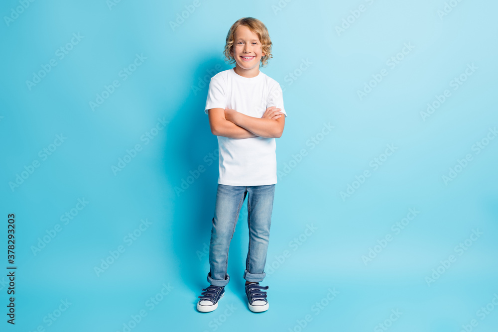 Full length photo of little kid crossed arms wavy hairdo look camera wear jeans white shirt sneakers isolated blue color background Full length photo of little kid crossed arms wavy hairdo look camera wear jeans white shirt sneakers isolated blue color background