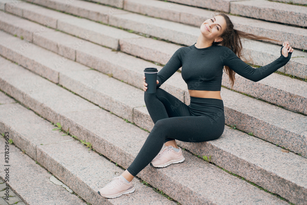 a beautiful young woman in sports clothes is sitting on the steps in the center of the Park, drinking coffee from the cafeteria in a paper Cup. he smiles and looks at the camera