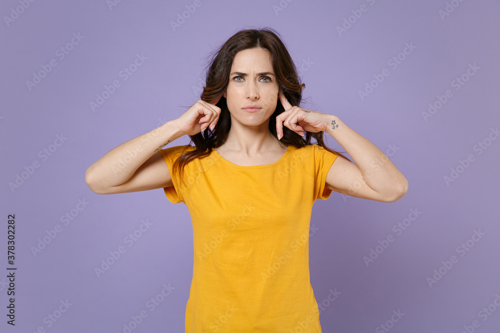 Displeased upset dissatisfied young brunette woman 20s wearing basic yellow t-shirt posing stand covering ears with fingers looking camera isolated on pastel violet colour background, studio portrait. Displeased upset dissatisfied young brunette woman 20s wearing basic yellow t-shirt posing stand covering ears with fingers looking camera isolated on pastel violet colour background, studio portrait.