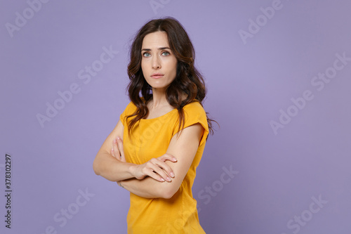 Side view of worried dissatisfied young brunette woman 20s wearing basic yellow t-shirt posing stand holding hands crossed looking camera isolated on pastel violet colour background, studio portrait.