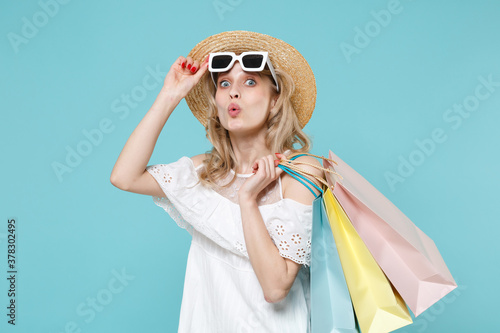 Shocked young blonde woman 20s wearing white summer dress hat eyeglasses hold package bag with purchases after shopping looking camera isolated on blue turquoise colour background studio portrait.