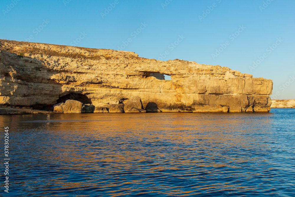 Seascape, a large rock in the form of a crocodile's head against the background of a clear blue sky and blue sea, with an orange reflection in the water