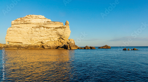 Seascape, a large rock in the form of a ship against the background of a clear blue sky and blue sea, with an orange reflection in the water