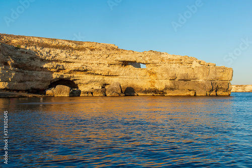Seascape, a large rock in the form of a crocodile's head against the background of a clear blue sky and blue sea, with an orange reflection in the water