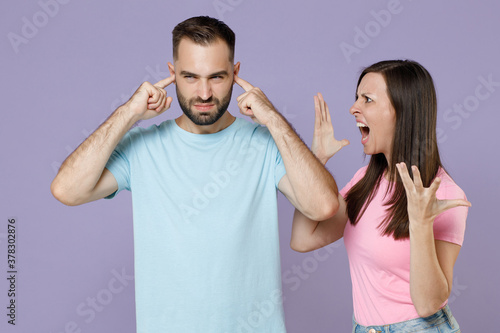 Displeased angry young couple two friends man woman 20s in blue pink t-shirts covering ears with fingers screaming swearing spreading hands isolated on pastel violet color background studio portrait.