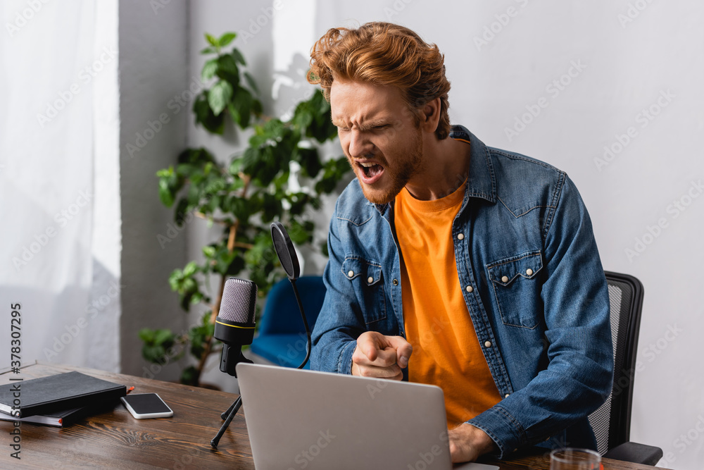angry broadcaster in denim shirt pointing with finger while screaming in microphone near laptop angry broadcaster in denim shirt pointing with finger while screaming in microphone near laptop