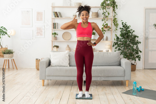 African Girl Standing On Weight Scales Gesturing Thumbs Up Indoors
