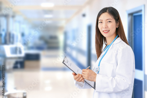 Smiling Asian female doctor wearing lab coat and stethoscope as she writes a document in her folder. In the blurred background you can see the hospital ward with medical equipment and patient rooms