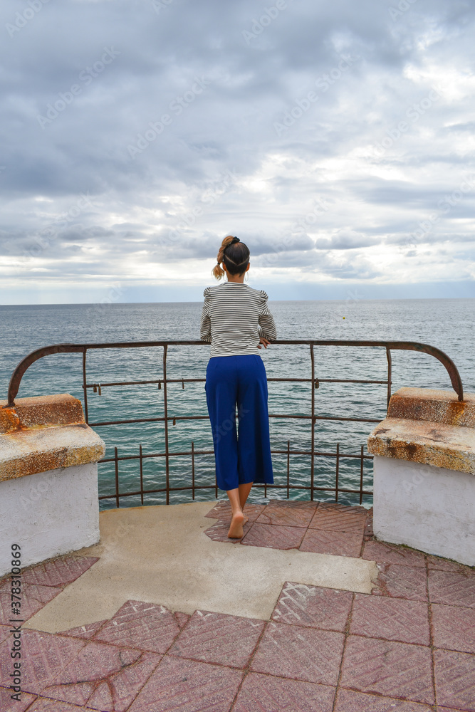 Woman photographed from the back and with bare feet, looking out to sea on a stormy day.