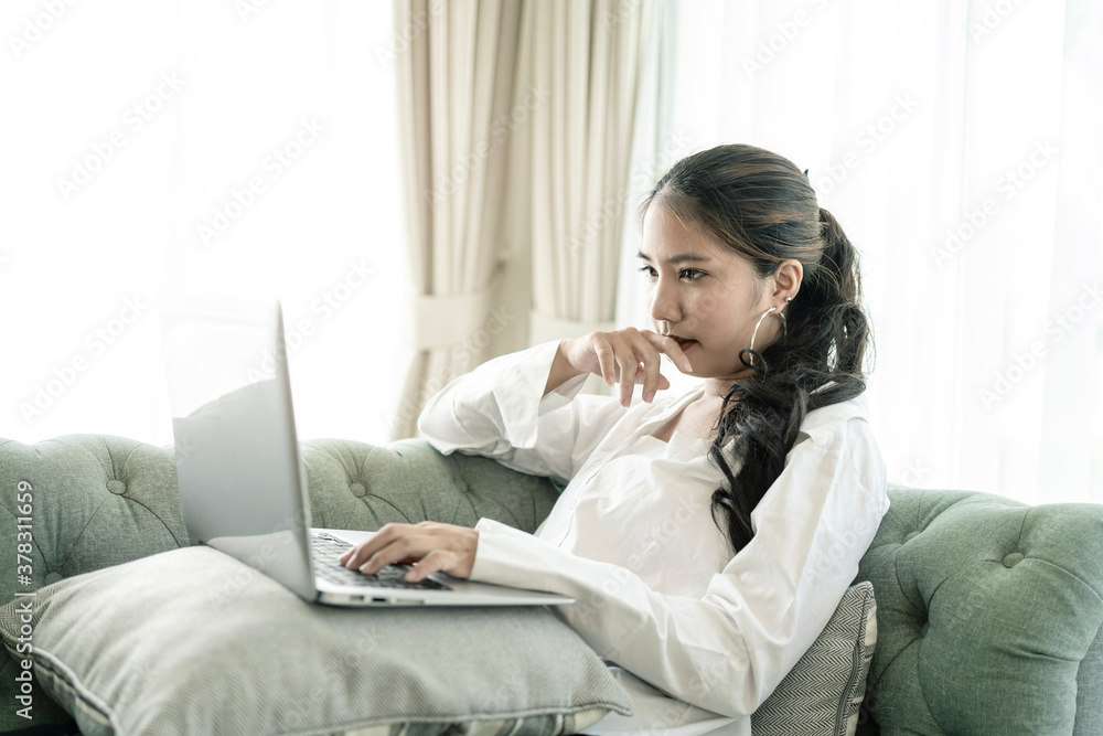 Happy casual beautiful Asian woman working on a laptop sitting on the couch in the house. Happy casual beautiful Asian woman working on a laptop sitting on the couch in the house.