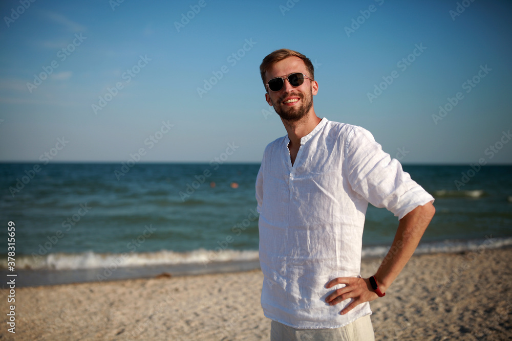 Young smiling man stands on beach against sea and sky background.