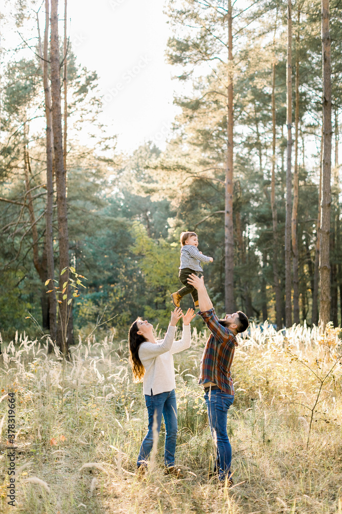Outdoor portrait of happy young parents, having fun and lifting up their little cute baby son, during walk in autumn forest at sunny day