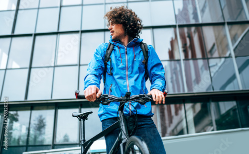 Fotografia Young man in blue raincoat standing with his bike after cycling in the street on a rainy day