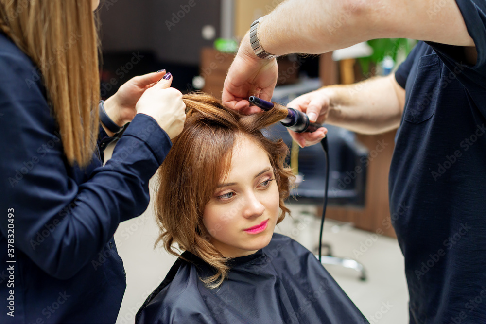 Professional hairdresser twists curls of long light brown hair of woman with curling iron in beauty salon. Hairdressing procedures Professional hairdresser twists curls of long light brown hair of woman with curling iron in beauty salon. Hairdressing procedures