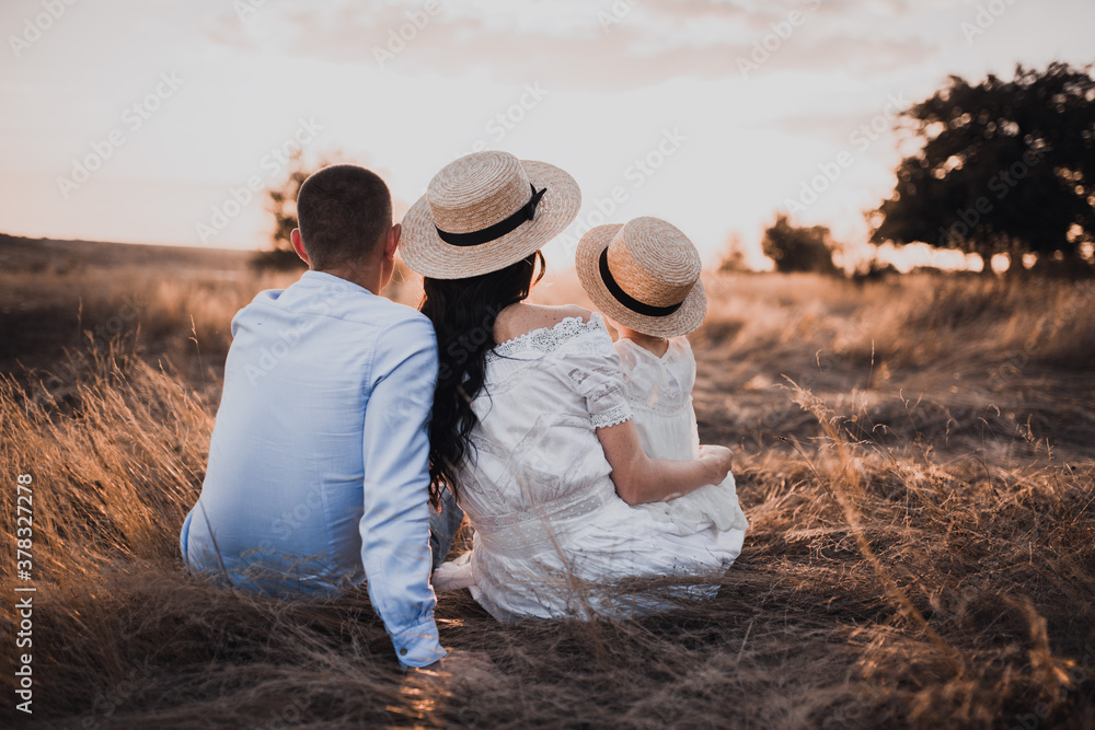 father, brunette mother and little daughter are sitting on the dry grass in white dresses. On the Sunset. in hats boater with his back. family