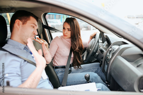 Male auto instructor takes exam in young woman. Female student look at teacher with confusion and misunderstanding. Guy show hands and look down. Daylight.