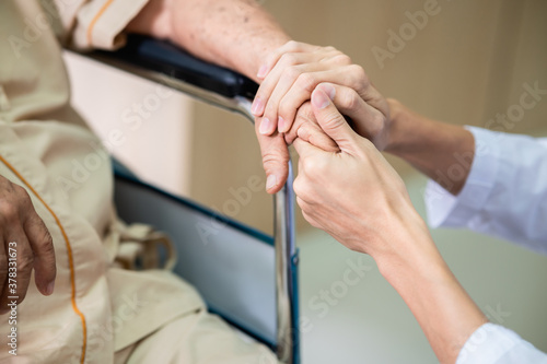 Close up hands of medical doctor carefully holding senior patient's hands in comfort during recovery in hospital. Asian doctor consoling elderly patients hand while sitting in wheelchair. 