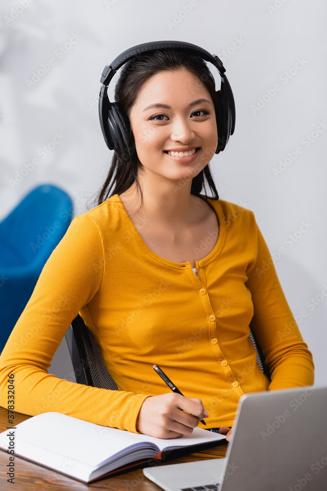brunette asian freelancer in wireless headphones looking at camera while holding pen near notebook brunette asian freelancer in wireless headphones looking at camera while holding pen near notebook