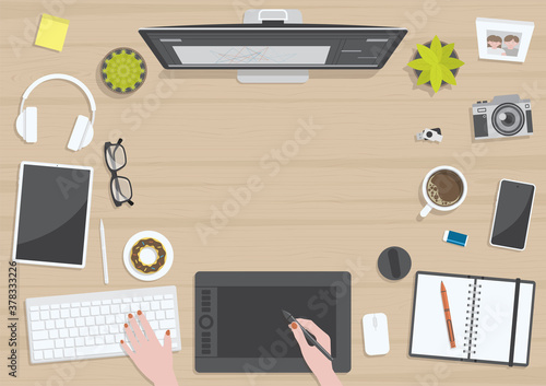 Wood textured table from top view with hands of woman drawing on digital graphic tablet with monitor, camera, tablet, headphones and gadgets, design for work from home concept