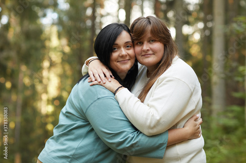 Portrait of two young lesbians embracing each other and smiling at camera while standing in the forest