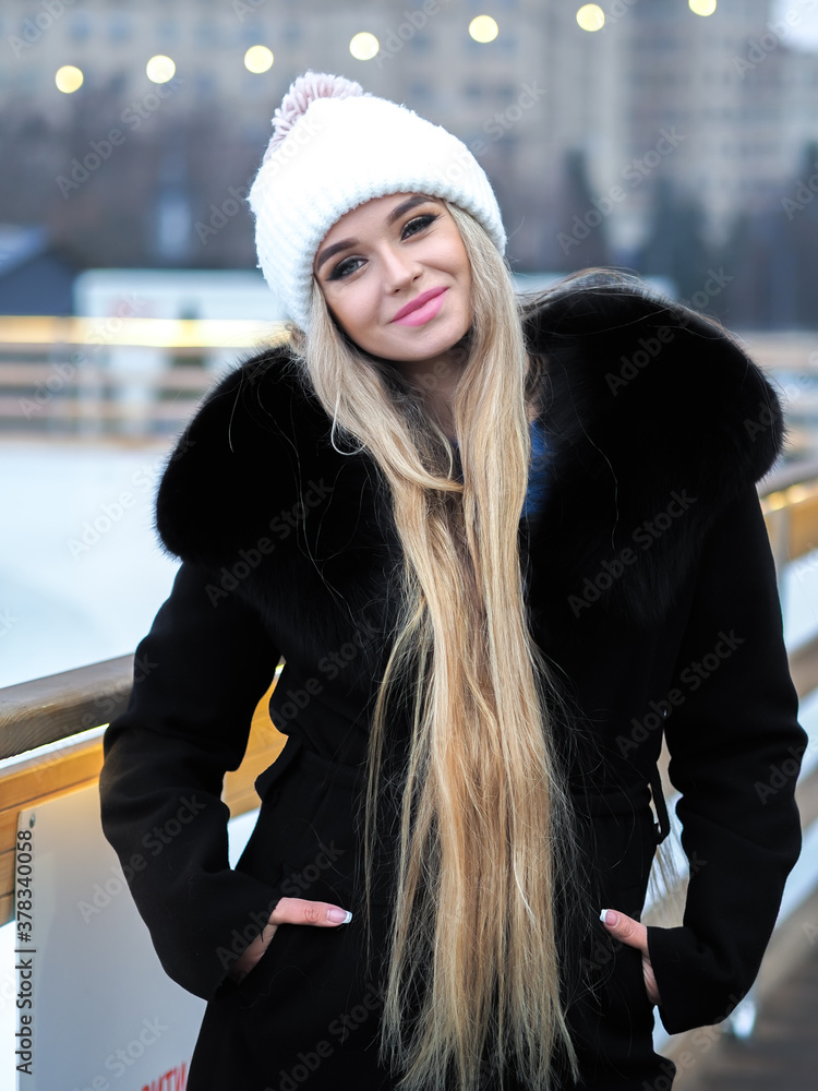 Blonde girl posing at the winter city in a black fur coat and a white hat