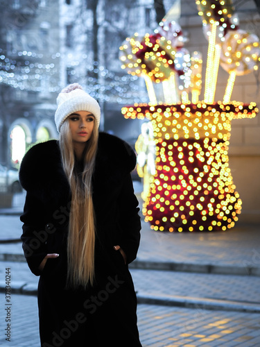 Blonde girl posing at the winter city in a black fur coat and a white hat