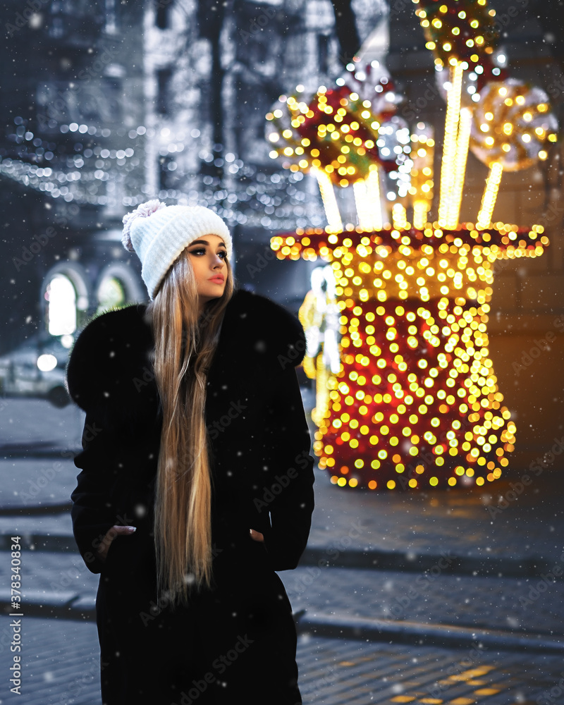Blonde girl posing at the winter city in a black fur coat and a white hat