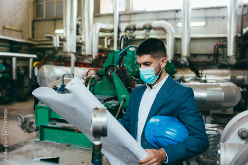 businessman holding blueprints in industry plant, wearing face mask, covid