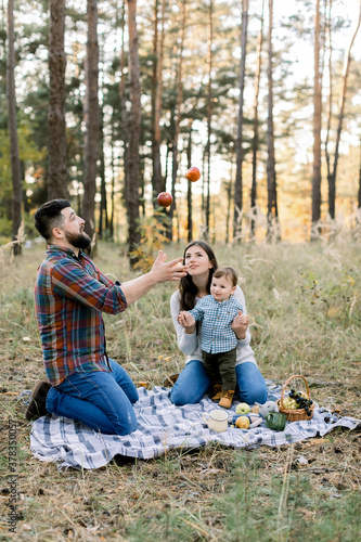 Family, fun and leisure concept. Portrait of happy family sitting on checkered plaid in autumn forest. Pretty mom and little baby boy having fun and looking at handsome father, juggling apples © sofiko14