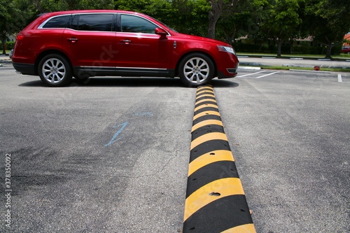 Red Minivan Driving Up to and Just Connecting with Yellow and Black Striped Speed Bump in Parking Lot with Diagonal Striped Spaces and Trees in the Background, Mid-Day photo