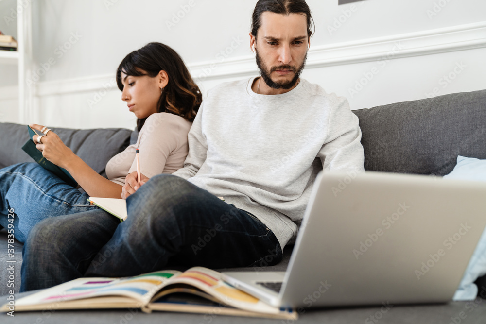 Fototapeta premium Image of serious beautiful couple reading book and working with laptop