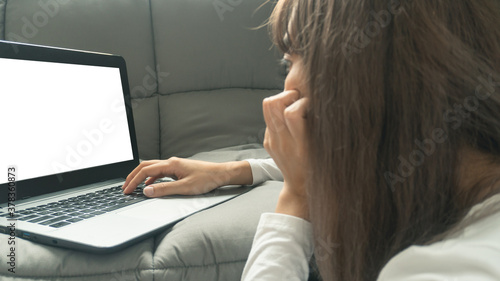 Woman rested her chin in her hands in the foreground and working or learning on laptop sitting on the grey sofa while working from home in quarantine for Covid-19 outbreak. Work from home concept.