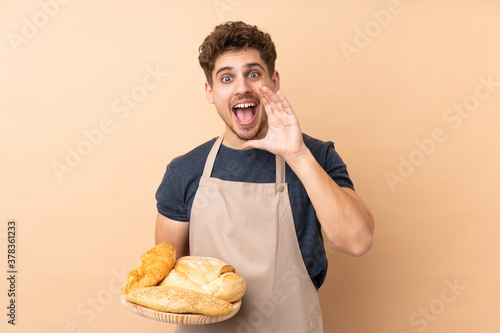 Male baker holding a table with several breads isolated on beige background shouting with mouth wide open