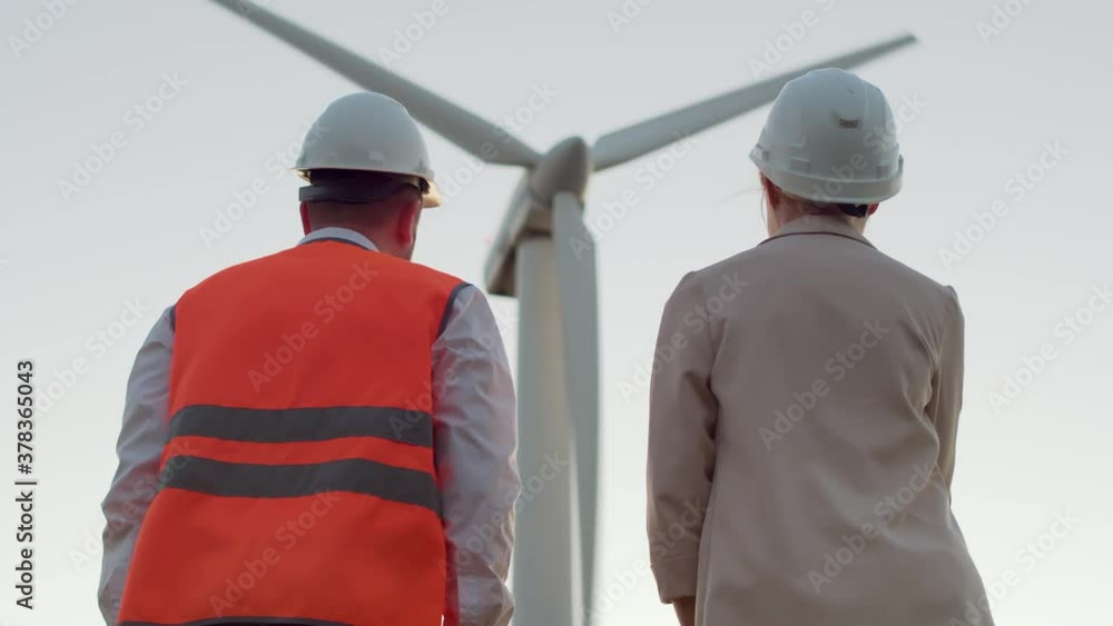 View from the back of the engineer and the girl in white helmets looking at the windmill, turn to each other and smiling shake hands View from the back of the engineer and the girl in white helmets looking at the windmill, turn to each other and smiling shake hands