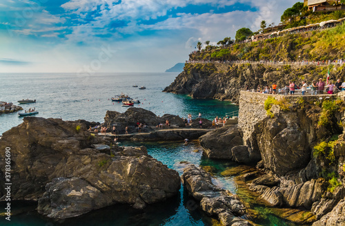 Beautiful panoramic landscape view of the coastal area in Manarola near the marina on a sunny day with blue sky. Tourists are following the trail up the cliff to the popular Manarola lookout point.